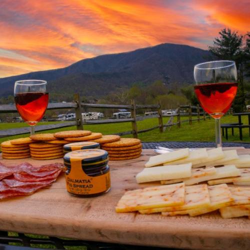 A scenic sunset view with two glasses of wine, cheese, crackers, and salami on a wooden platter.