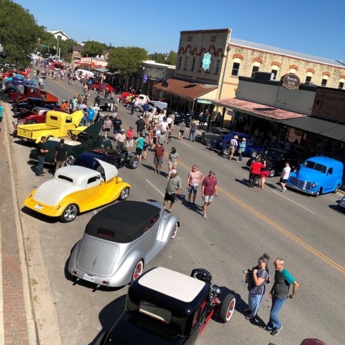 A vibrant street scene with classic cars and crowds of people enjoying a car show under a clear blue sky.