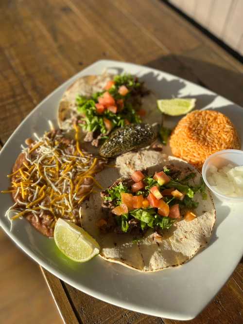 A plate of tacos with beef, lettuce, and tomatoes, served with rice, refried beans, and lime wedges.