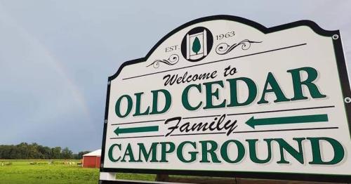 Sign for Old Cedar Family Campground, established in 1963, with a rainbow in the background and green fields.