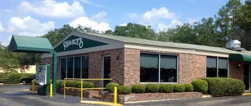 A brick restaurant building with large windows and a green awning, surrounded by neatly trimmed bushes and trees.