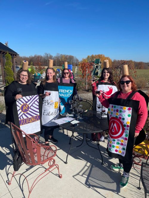 Five women in creative costumes holding signs for different wineries, standing outdoors with a clear blue sky.