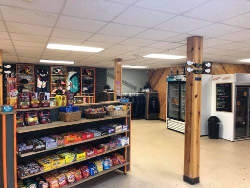 Interior of a convenience store with shelves of snacks, drinks, and a cooler, featuring wooden pillars and a chalkboard menu.
