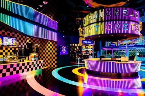 Brightly lit arcade entrance with colorful neon lights, ticket counter, and checkered walls. Fun, vibrant atmosphere.