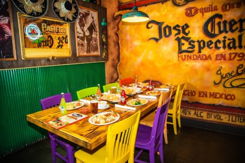 A vibrant restaurant table set with colorful chairs, plates of food, and drinks against a lively mural backdrop.