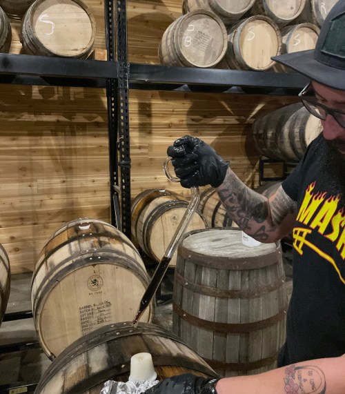 A person in a black shirt and gloves uses a siphon to draw liquid from a wooden barrel in a storage area.