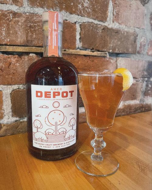 A bottle of Amer Depot liqueur next to a glass of cocktail garnished with a lemon twist, set against a brick wall.