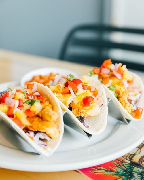 Three tacos filled with crispy fish, topped with diced tomatoes, onions, and cilantro, served on a white plate.