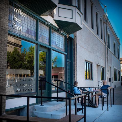 A café exterior with a sign for coffee, beer, and food; two people sit at a table outside enjoying drinks.