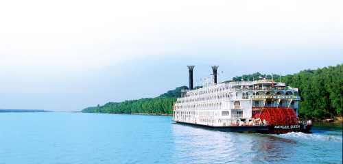 A large riverboat with paddle wheels sails along a calm blue river, surrounded by lush green trees.