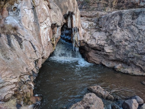 A rocky landscape featuring a waterfall flowing through a cave, with icicles hanging from the stone above.