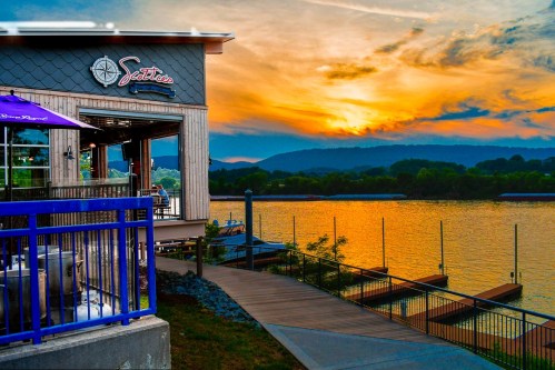 Sunset over a lake with a restaurant on the shore, featuring a deck and mountains in the background.