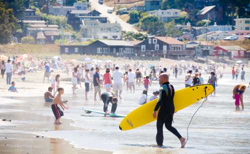 A surfer in a wetsuit carries a yellow surfboard along a crowded beach with people enjoying the sun and water.