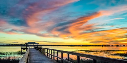 A wooden pier extends into a calm lake at sunset, with vibrant clouds reflecting in the water.