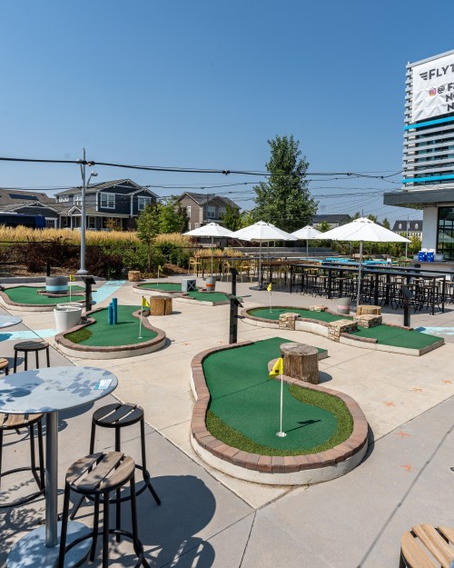 A sunny outdoor mini-golf course with green turf, wooden obstacles, and seating areas in the background.