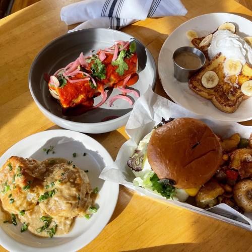 A table with four dishes: enchiladas, French toast with bananas, a burger with salad, and a side of potatoes.