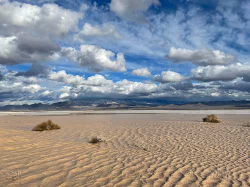 A vast desert landscape with rippled sand, sparse vegetation, and dramatic clouds against a distant mountain range.