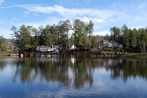 A serene lake reflects houses and tall pine trees under a clear blue sky.