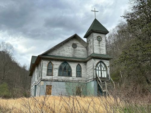 An old, weathered church with a steeple and cross, surrounded by overgrown grass and trees under a cloudy sky.
