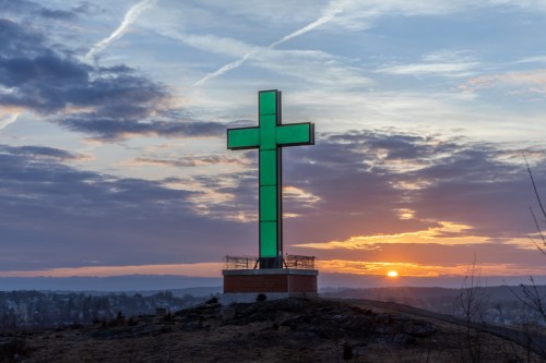 A large illuminated cross stands on a hill against a colorful sunset and cloudy sky.