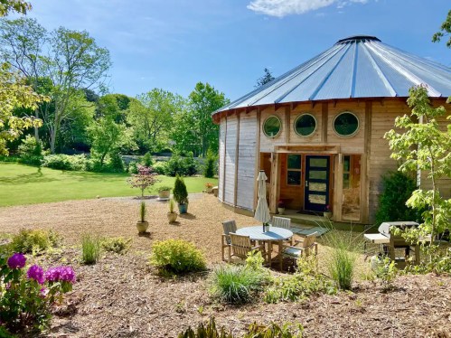A round wooden building with a metal roof, surrounded by greenery and flowers, featuring outdoor seating.