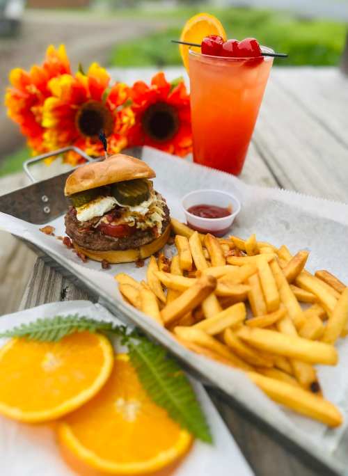 A delicious burger with fries, garnished with pickles and a fried egg, alongside a colorful drink and orange slices.