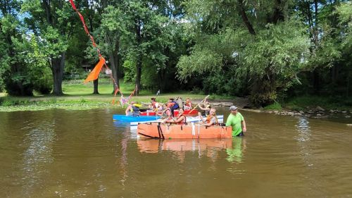 A group of people in colorful boats paddles on a calm river surrounded by trees on a sunny day.
