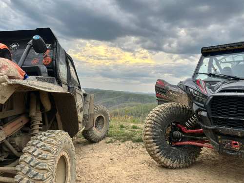 Two off-road vehicles parked on a dirt trail, overlooking a scenic landscape under a cloudy sky.