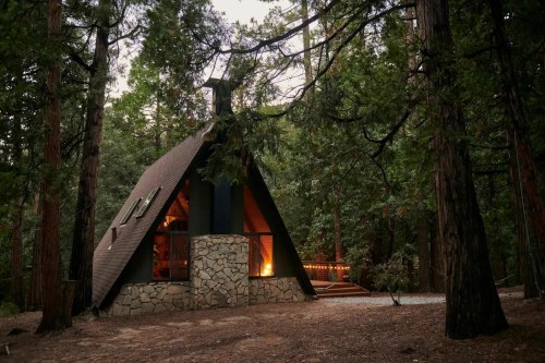 A cozy A-frame cabin nestled among tall trees, with a warm glow from the fireplace visible through large windows.