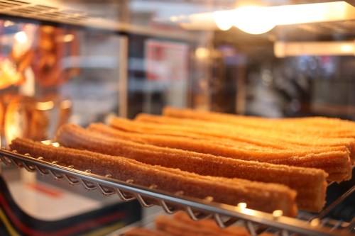 A close-up of golden-brown churros displayed in a glass case, illuminated by warm light.