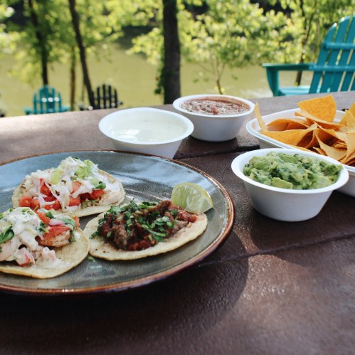 Two tacos with toppings, guacamole, salsa, sour cream, and tortilla chips on a table by a scenic outdoor setting.