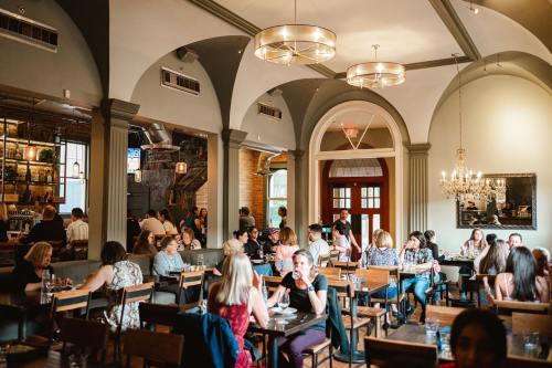 A bustling restaurant interior with diners at wooden tables, high ceilings, and elegant lighting fixtures.