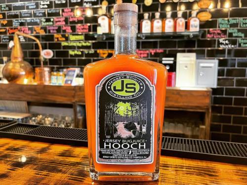A bottle of "Jersey Bug Juice Hooch" on a bar, featuring a colorful label with a forest scene and the distillery logo.