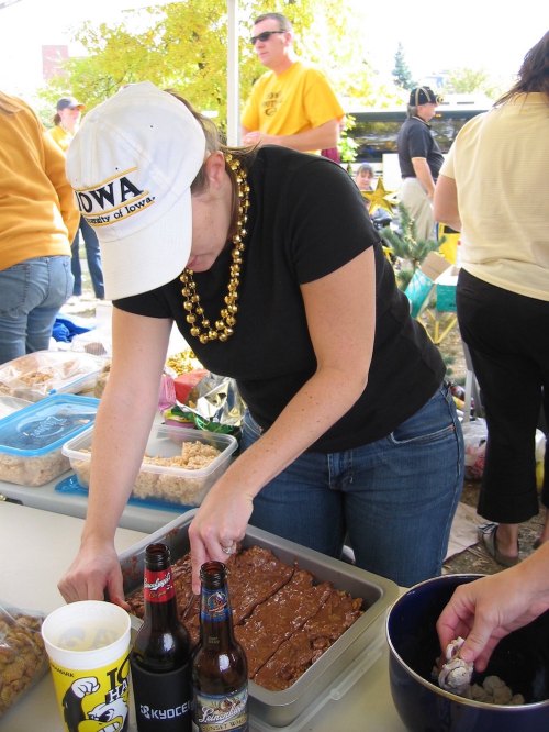 A person in a black shirt and cap prepares food at a gathering, with drinks and snacks visible on the table.