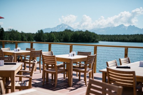Outdoor dining area with wooden tables overlooking a lake and mountains under a clear blue sky.