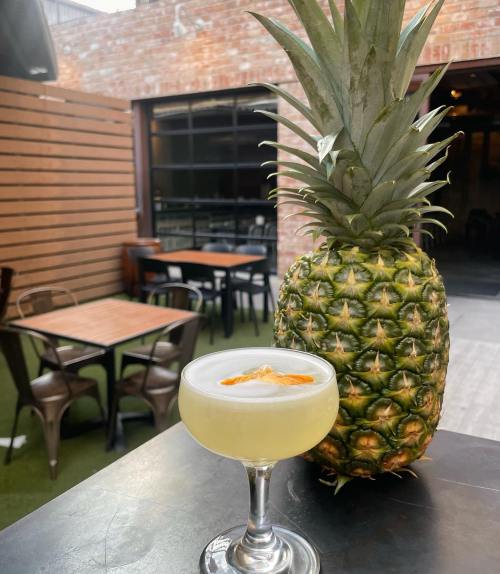 A cocktail in a coupe glass garnished with orange sits next to a fresh pineapple in a cozy outdoor setting.