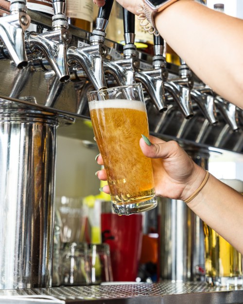 A hand pours a golden beer from a tap into a clear glass at a bar.