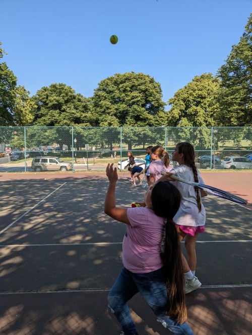 Children playing tennis on a court, with one girl preparing to hit a ball while others watch. Sunny day with trees in background.