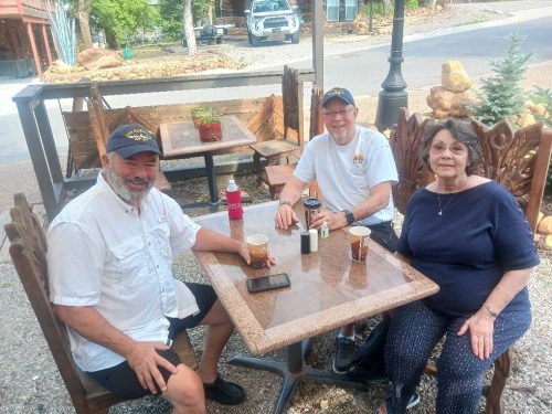 Three people sit at an outdoor table, enjoying drinks and smiling, with a rustic setting in the background.