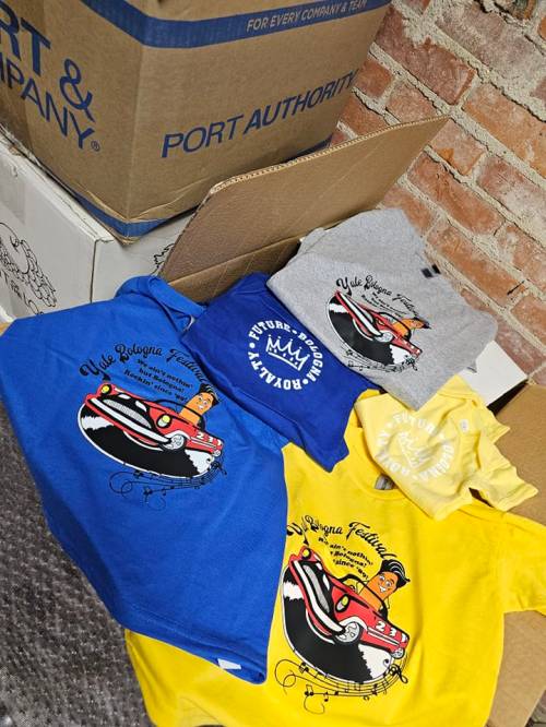 A collection of colorful t-shirts featuring a cartoon car design, stacked on cardboard boxes against a brick wall.