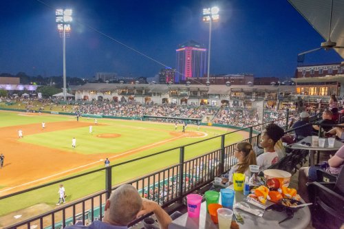 A lively baseball game at night, with fans enjoying drinks and snacks from a balcony overlooking the field.