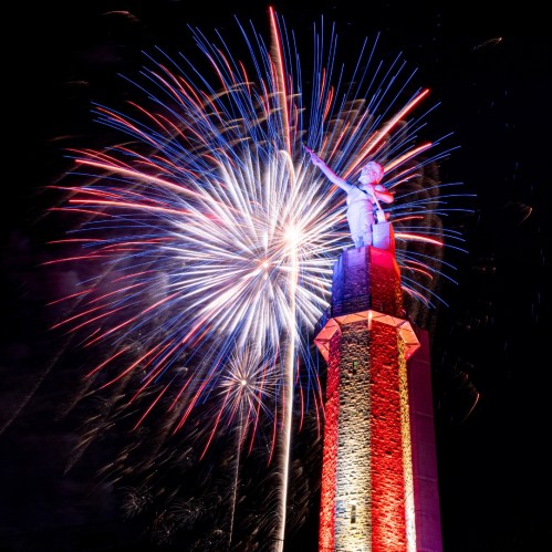 Fireworks burst behind a tall statue on a stone tower, illuminated in red and blue against a dark sky.