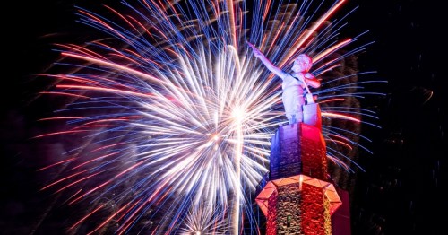 A statue atop a monument with colorful fireworks bursting in the night sky behind it.
