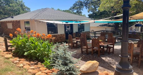 A cozy outdoor seating area with wooden chairs, vibrant flowers, and a charming building under a clear blue sky.
