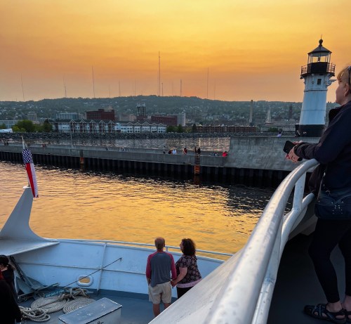 A sunset view from a boat, with people on deck and a lighthouse in the background along the shoreline.