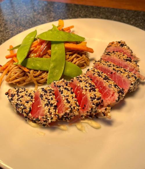 Seared sesame-crusted tuna slices served with stir-fried noodles and vegetables on a white plate.