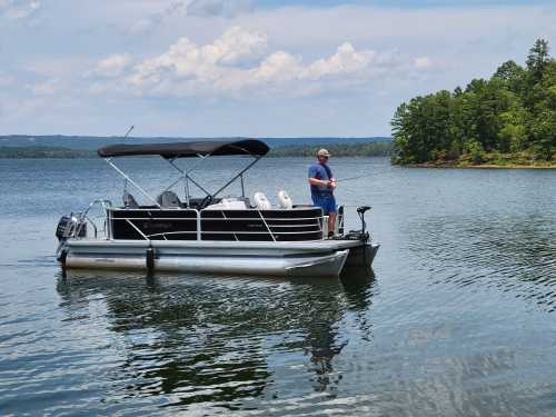 A man stands on a pontoon boat in calm waters, surrounded by trees and a cloudy sky.