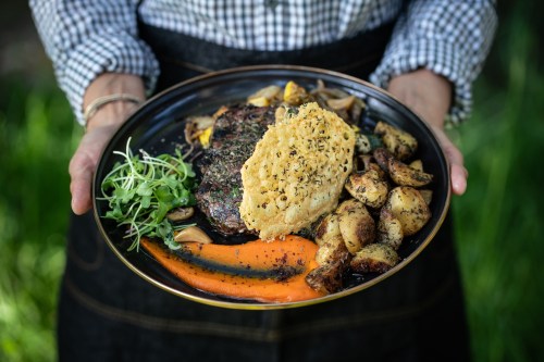 A person holds a plate with steak, roasted potatoes, carrots, and a herb crust, set against a green background.