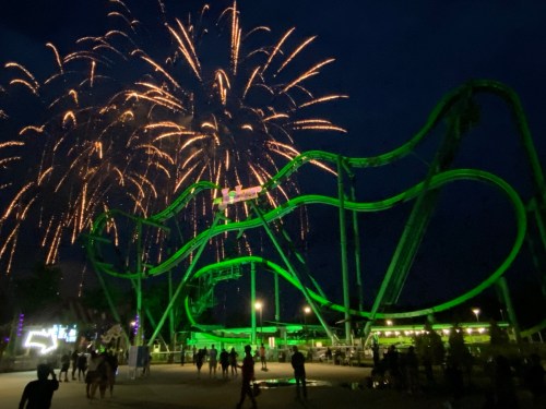 A roller coaster illuminated in green with fireworks bursting in the night sky above a crowd.