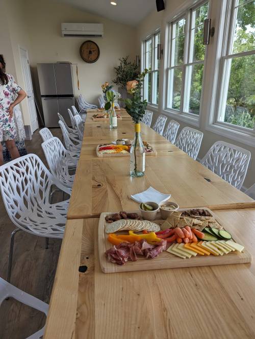A long wooden table set with a charcuterie board, drinks, and flowers, surrounded by white chairs in a bright room.